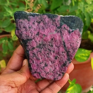 May include: A hand holds a rough gemstone, showcasing a striking contrast of pink and black. The stone's surface is textured, with pink areas speckled with darker spots. The top edge of the stone is a solid black.