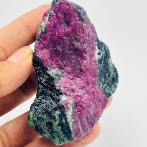 May include: A raw ruby gemstone, displaying a rich pink and red colour, with dark green and black mineral inclusions. The natural stone is held in a hand, highlighting its irregular shape and textured surface.