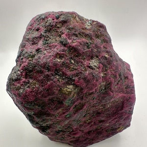 May include: A large, rough-hewn ruby gemstone with a deep red hue, accented by dark green and black patches. The surface has a textured appearance, showcasing natural mineral formations. The gemstone is displayed against a white background.