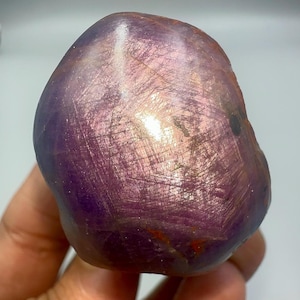Może przedstawiać: Wypolerowany, zaokrąglony kamień szlachetny o głębokim fioletowym odcieniu, wykazujący błyszczącą, opalizującą powierzchnię. Tekstura kamienia ukazuje subtelne prążki i wariacje kolorystyczne, z nutami różu i złota. Ogólny kształt jest gładki i organiczny.