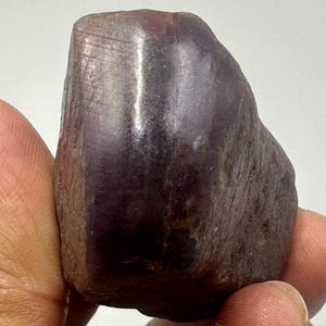 May include: A natural, unpolished gemstone in a deep purple colour. The stone has a rough, textured surface and a rounded shape. The colour varies from dark purple to a lighter shade, with some areas appearing almost brown. The gemstone is held in a hand.