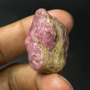 Può includere: Una gemma di rubino grezza e non tagliata. Il rubino ha un colore rosa intenso con aree beige e marroni. La superficie è irregolare e strutturata, con un aspetto naturale e non lucido. La gemma è tenuta in mano su uno sfondo scuro.
