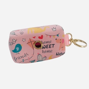 Op de afbeelding: Roze hondenpoepzakjesdispenser met een goudkleurige clip. De dispenser is versierd met cartoonillustraties van een vogel, een hond, harten en de woorden "home SWEET home" en "friends".