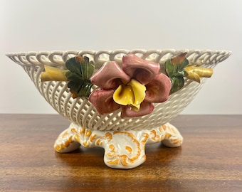Op de afbeelding: Een witte keramische kom met een geweven ontwerp en drie poten. De kom is versierd met een roze en gele bloem met groene bladeren.