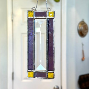 May include: A rectangular stained glass piece with a clear, faceted center and a purple border. Yellow squares accent the top and bottom. A silver metal hanger is attached to the top, allowing it to be suspended.