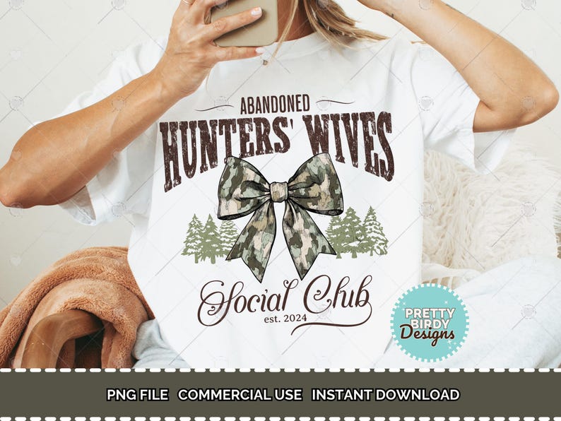以下が含まれることがあります： 白いTシャツに、茶色の文字で「Abandoned Hunters' Wives Social Club est. 2024」と書いてあります。茶色と緑の迷彩柄のリボンがテキストの上に中央に配置されています。リボンの両側に小さな緑の木が2本あります。