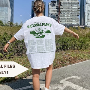 May include: White t-shirt with a green graphic print of mountains and a sun. The text "United States National Parks" is printed above the graphic. The text "The Heart of America's Wildlands" is printed below the graphic.