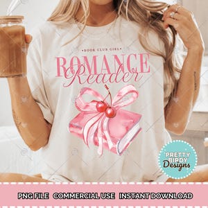 以下が含まれることがあります： ピンク色の「ROMANCE Reader」の文字が入ったクリーム色のTシャツ。ピンク色の本にさくらんぼが乗った水彩画イラストが特徴です。Tシャツには、メインテキストの上に「BOOK CLUB GIRL」の文字も入っています。
