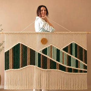 May include: A large macrame wall hanging featuring a mountain landscape design in shades of dark green, light green, and brown. The piece includes a circular mustard yellow sun element and is suspended from a horizontal wooden dowel with fringe.
