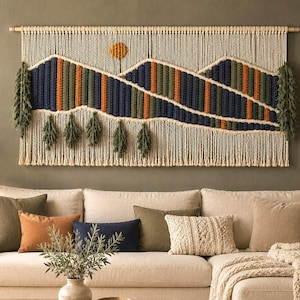 May include: A large rectangular macrame wall hanging featuring a mountain landscape design in cream, navy blue, olive green, and burnt orange cord. The piece includes a circular orange sun, textured mountain peaks, and hanging fringe tassels.