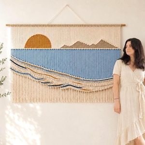 Puede incluir: Un gran tapiz de macramé rectangular que representa una puesta de sol sobre el océano. La obra de arte presenta un sol dorado, agua azul y flecos beige, con montañas marrones en el fondo. Está suspendido de una barra de madera.