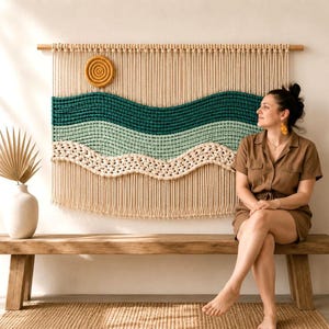 Puede incluir: Un gran tapiz de macramé para pared con un diseño de paisaje que incluye un sol amarillo mostaza, patrones de ondas en cuerda de color verde azulado oscuro, verde claro y crema, colgado de un listón de madera con flecos verticales.