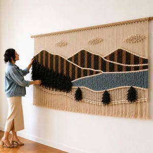 May include: A large macrame wall hanging featuring a landscape design with beige, dark green, brown, and blue fiber cords. The piece depicts mountains, clouds, and trees, suspended from a wooden rod with a long fringe along the bottom edge.