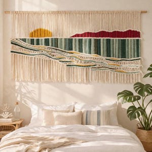 Könnte beinhalten: Ein großer, gewebter Wandbehang mit Landschaftsdesign. Das Kunstwerk zeigt eine gelbe Sonne, rote Berge und grün-blaue Strukturelemente. Das Stück ist an einer Holzstange aufgehängt und hat lange, cremefarbene Fransen.