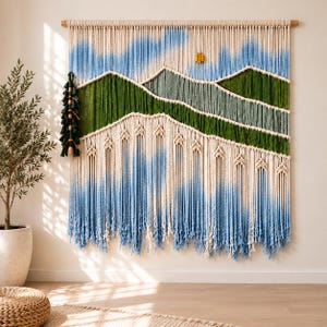 May include: A large macrame wall hanging featuring a landscape design with cream, green, and blue dyed cotton cords. The piece depicts rolling hills and a sky, suspended from a wooden dowel, with a dark green tassel accent on the left side.