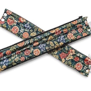 May include: Two long, rectangular fabric strap covers featuring a dark green background with a repeating pattern of orange slices, red pomegranate halves, and small blue berries. Each cover includes a black zipper, white snap buttons, and a white plastic clip.