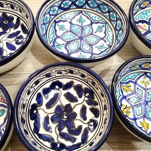 Może przedstawiać: Kolekcja ceramicznych misek z białymi wnętrzami i ciemnoniebieskimi obwódkami. Każda miska ma unikalny, ręcznie malowany kwiatowy wzór w odcieniach niebieskiego, turkusowego i akcentach czerwieni i żółci. Miski ułożone są na jasnej drewnianej powierzchni.