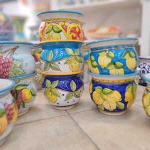 Może przedstawiać: Kolekcja ręcznie malowanych ceramicznych donic z żywymi wzorami. Donice zdobią kolorowe wzory owoców i kwiatów w odcieniach żółci, błękitu i czerwieni. Donice są ułożone w stosy i ustawione na kafelkowej powierzchni.