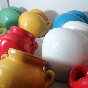 Könnte beinhalten: Eine Sammlung farbenfroher Keramikvasen in verschiedenen Farbtönen, darunter Rot, Gelb, Grün, Blau und Weiß. Jede Vase hat eine abgerundete Form mit einem kleinen Henkel. Die Vasen sind eng beieinander angeordnet und bilden eine lebendige Darstellung.