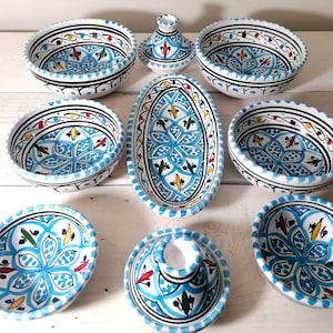 Op de afbeelding: Een verzameling handbeschilderde keramische serveerschalen met een levendig blauw, wit, zwart, rood en geel bloemmotief. De set bevat kommen, een ovale schaal en een kleine container met deksel, perfect voor het serveren van voedsel.