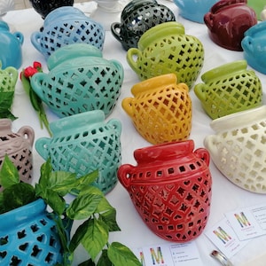 Può includere: Una collezione di vasi in ceramica colorati con intricati disegni a traforo. I vasi sono in vari colori, tra cui blu, verde, giallo e rosso. Ogni vaso ha due manici e un motivo decorativo. Alcuni vasi contengono piante.