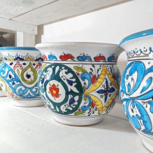 May include: A row of ceramic pots with colorful, hand-painted designs. The pots feature white bases, wide rims, and intricate patterns in blue, green, yellow, and red. The pots are displayed on a white shelf, showcasing their decorative appeal.