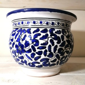 Puede incluir: Maceta de cerámica blanca con diseño floral azul. La maceta tiene un cuerpo ancho y redondeado y un borde acampanado con un borde azul oscuro. El patrón floral es en tonos de azul y gris.