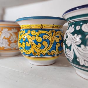 Peut inclure: Trois pots en céramique avec des motifs floraux colorés. Les pots ont une forme arrondie avec une large ouverture et un bord légèrement évasé. Le pot central présente un motif bleu et jaune, tandis que les autres ont des motifs orange et vert.