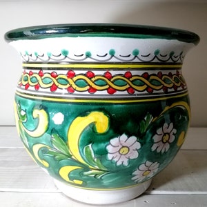 Peut inclure: Un pot de fleurs en céramique peint à la main, de forme large et arrondie. Le pot a un bord vert foncé et une base blanche. Le corps est décoré de motifs floraux verts, jaunes et rouges et de fleurs blanches en forme de marguerites.