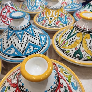 Pode incluir: Uma coleção de tagines de cerâmica pintados à mão e coloridos. Cada panela apresenta desenhos intrincados em azul, amarelo, vermelho e verde, com uma base branca e uma borda amarela. As tampas têm formato cônico e uma pequena abertura superior.