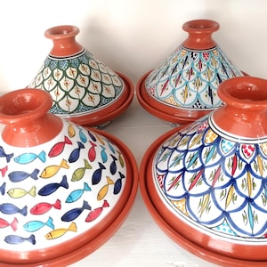 Pode incluir: Quatro tagines de terracota com desenhos pintados em branco, vermelho, azul, amarelo e verde. Os tagines têm tampas cónicas e bases redondas. Dois tagines apresentam padrões de peixes, enquanto os outros têm padrões geométricos e florais.