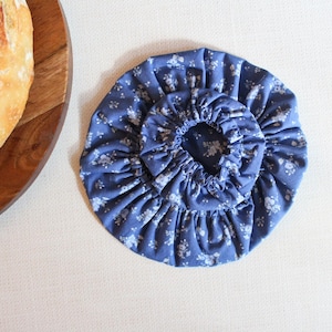 May include: A blue floral fabric bread cover with an elastic edge. The cover is designed to fit over a loaf of bread. A wooden breadboard and a loaf of bread are partially visible in the image.