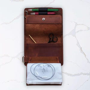 Puede incluir: Estuche abierto de cuero marrón para agujas de tejer con varias herramientas. El estuche contiene agujas de tejer verdes y rojas, tijeras negras, un ganchillo de madera y una bolsa de plástico transparente con una aguja circular. El estuche está abierto y listo para usar.