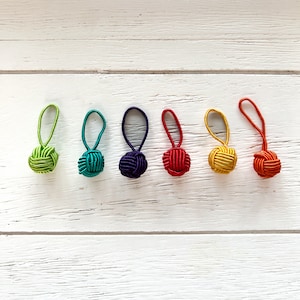 Op de afbeelding: Zes kleurrijke decoratieve kwastjes met knoop, in een rij. De kwastjes zijn groen, turquoise, paars, rood, geel en oranje. Elke kwast heeft een lus en een geknoopte bal, tegen een witte houten achtergrond.