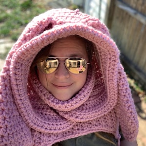 May include: A pink crocheted hooded scarf with a thick, textured design. The scarf is worn by a person wearing sunglasses.