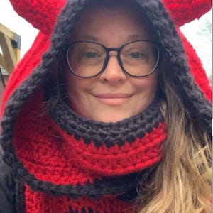 May include: A red and black crocheted hooded scarf with horns. The scarf is worn by a person with long blonde hair and glasses.