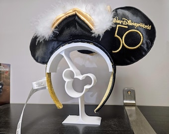 May include: White plastic headband holder with a Mickey Mouse silhouette. A black headband with gold trim and white feathers is displayed on the holder. The headband has the text "Walt Disney World 50" embroidered on it.