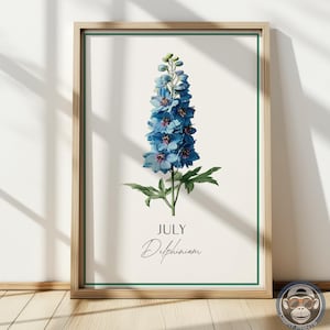K&ouml;nnte beinhalten: Ein gerahmter botanischer Druck mit einer leuchtend blauen Delphiniumblume. Das Kunstwerk, vor einem cremefarbenen Hintergrund, enth&auml;lt den Text "JULY" und "Delphinium". Der Rahmen ist aus hellem Holz mit einem gr&uuml;nen Rand, geeignet f&uuml;r die Inneneinrichtung.