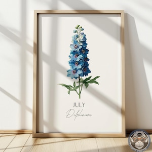 K&ouml;nnte beinhalten: Ein gerahmter botanischer Druck mit einer blauen Delphiniumblume. Das Kunstwerk enth&auml;lt das Wort "JULI" und den Namen der Blume in Schreibschrift. Der Druck ist vor einem cremefarbenen Hintergrund und in hellem Holz gerahmt.