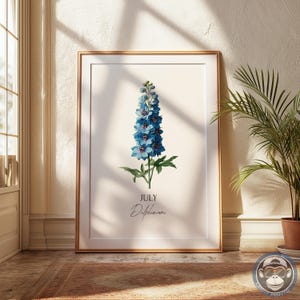 K&ouml;nnte beinhalten: Ein gerahmter botanischer Druck mit einer blauen Delphiniumblume. Der Druck enth&auml;lt das Wort "Juli" und den Namen der Blume, "Delphinium", in Schriftform. Das Kunstwerk ist in einem goldenen Rahmen gefasst und verleiht dem floralen Design einen Hauch von Raffinesse.