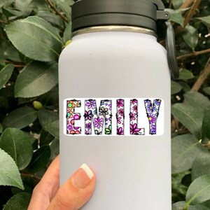 Pode incluir: Garrafa de &aacute;gua branca com tampa e al&ccedil;a pretas. O nome "Emily" est&aacute; escrito na garrafa com flores coloridas.