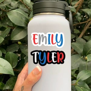 May include: White water bottle with a black lid and handle. The bottle has a sticker with the names "Emily Tyler" in red, blue, and black lettering.