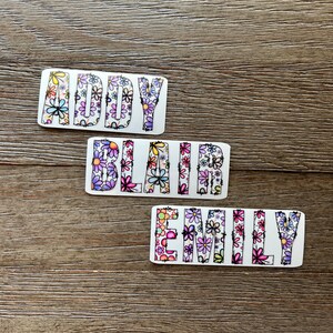 Pode incluir: Tr&ecirc;s adesivos brancos com os nomes "Addy", "Blair" e "Emily" escritos em letras pretas delineadas. As letras s&atilde;o preenchidas com um padr&atilde;o floral colorido.