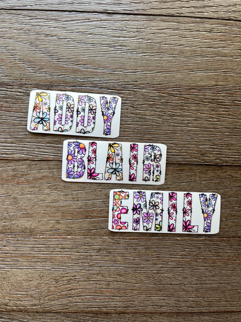 Pode incluir: Tr&ecirc;s adesivos brancos com os nomes "Addy", "Blair" e "Emily" escritos em letras pretas delineadas. As letras s&atilde;o preenchidas com um padr&atilde;o floral colorido.