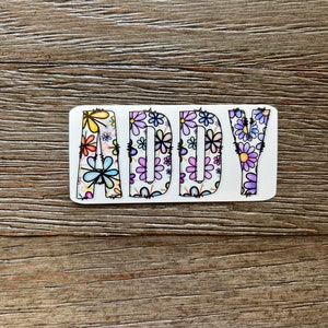 Pode incluir: Um adesivo branco com o nome "ADDY" escrito em letras pretas delineadas. As letras s&atilde;o preenchidas com um padr&atilde;o floral colorido.