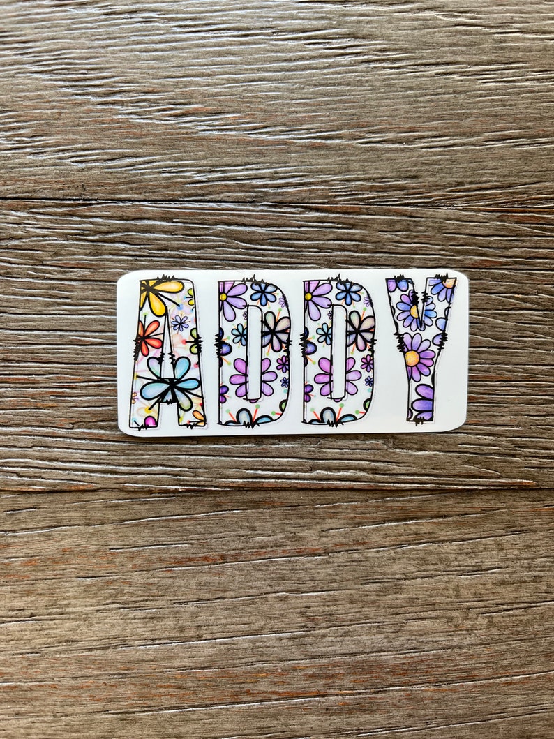 Pode incluir: Um adesivo branco com o nome "ADDY" escrito em letras pretas delineadas. As letras s&atilde;o preenchidas com um padr&atilde;o floral colorido.