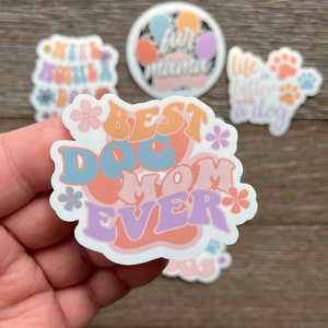 K&ouml;nnte beinhalten: Ein wei&szlig;er Aufkleber mit dem Text "Best Dog Mom Ever" in bunten, Retro-Lettern. Der Aufkleber zeigt ein Pfotenabdruck-Design und florale Akzente.
