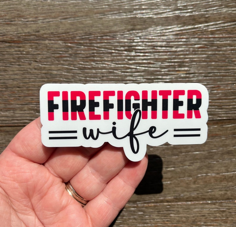 K&ouml;nnte beinhalten: Wei&szlig;er Aufkleber mit dem Text "FIREFIGHTER" in Rot und Schwarz mit einem d&uuml;nnen schwarzen Strich, der die W&ouml;rter trennt. Der Text "wife" ist in schwarzer Kursivschrift mit zwei schwarzen Linien auf jeder Seite des Wortes.
