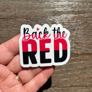 K&ouml;nnte beinhalten: Ein wei&szlig;er Aufkleber mit dem Text "Back the RED" in schwarzer und roter Schrift. Das Wort "RED" ist in zwei H&auml;lften geteilt, wobei die obere H&auml;lfte rot und die untere H&auml;lfte schwarz ist. Ein kleines rosa Herz befindet sich &uuml;ber dem Wort "Back".