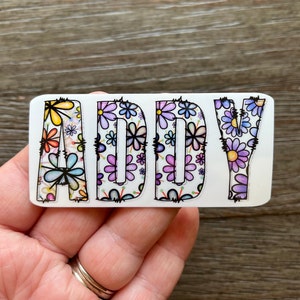 Pode incluir: Um adesivo branco com o nome "ADDY" escrito em letras com contorno preto. As letras s&atilde;o preenchidas com um padr&atilde;o floral colorido de flores roxas, azuis, amarelas e rosas.